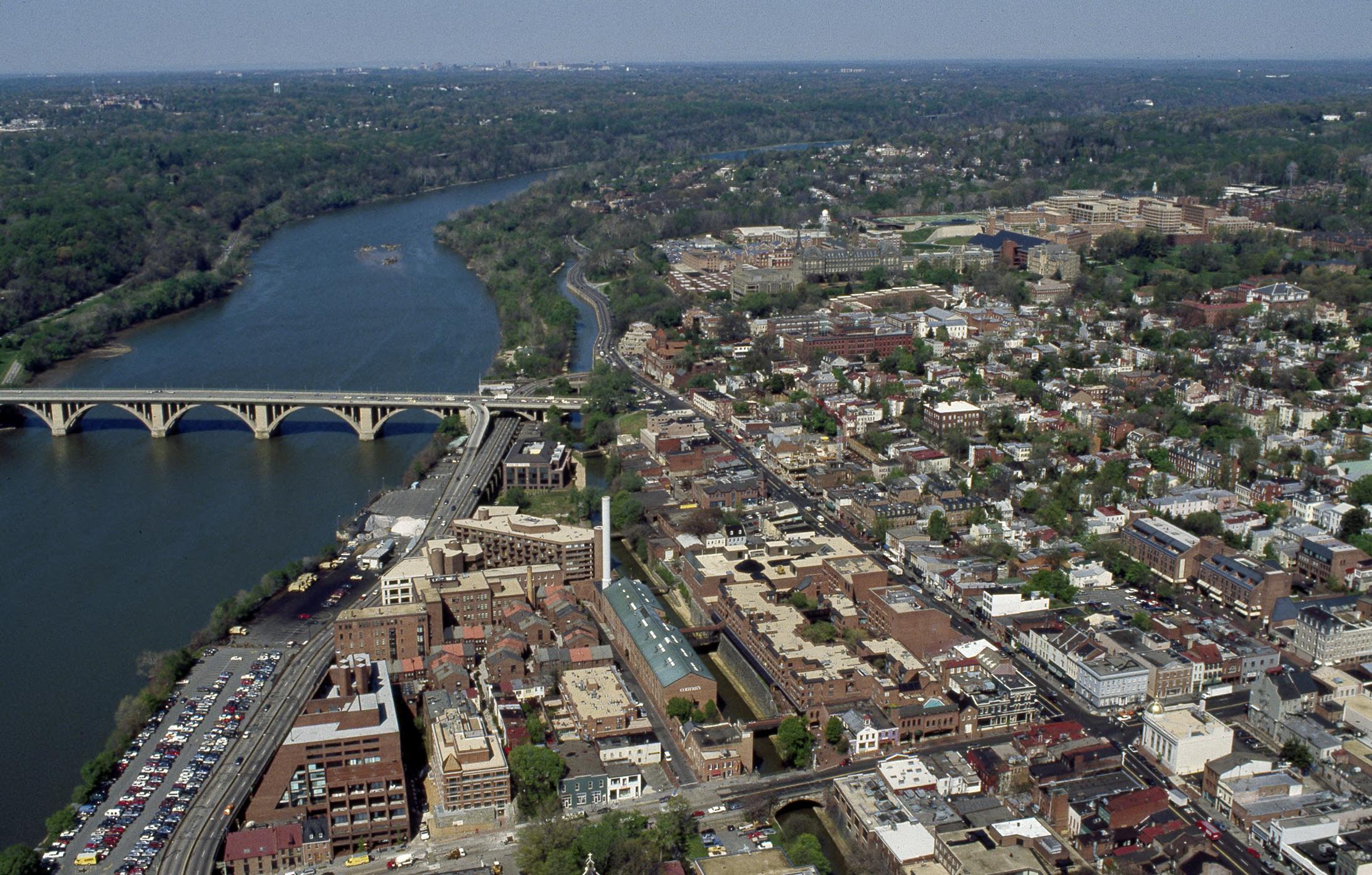 An arial view of Georgetown and the Potomac. On the right side of the photo are crowded buildings, to the left is the river and a bridge, and to the left of the river the land is covered with trees.