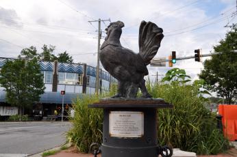 A photo of a life-size statue of Roscoe the Rooster at an intersection with a commemorative plaque on the base of the sculpture