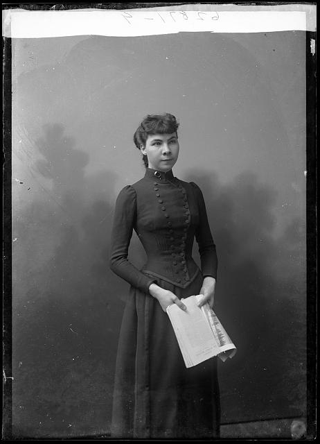 A 3/4 length photo of Madeline Pollard standing in a black dress with a newspaper in her hands