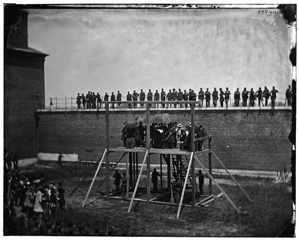 Photograph of the four conspirators on the scaffold with priests and soldiers holding umbrellas to block the sun. Guards stand on the wall behind them. Source: Library of Congress