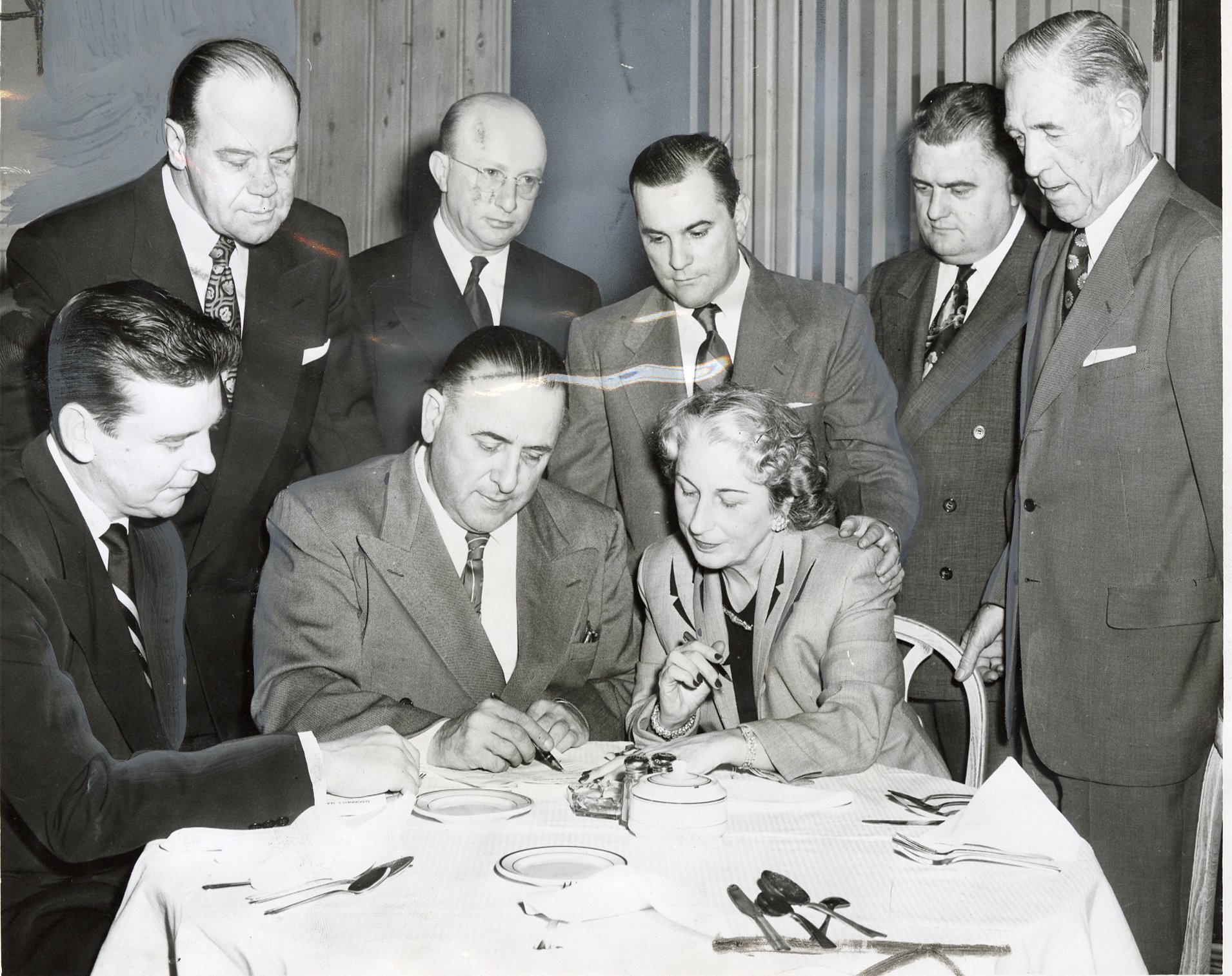 A group of people gather as Mrs. Florence Menendez signs the lease of Turner's Arena to Vince McMahon Sr., seated two spots from her right. Her husband, Gabe Menendez, stands over her.