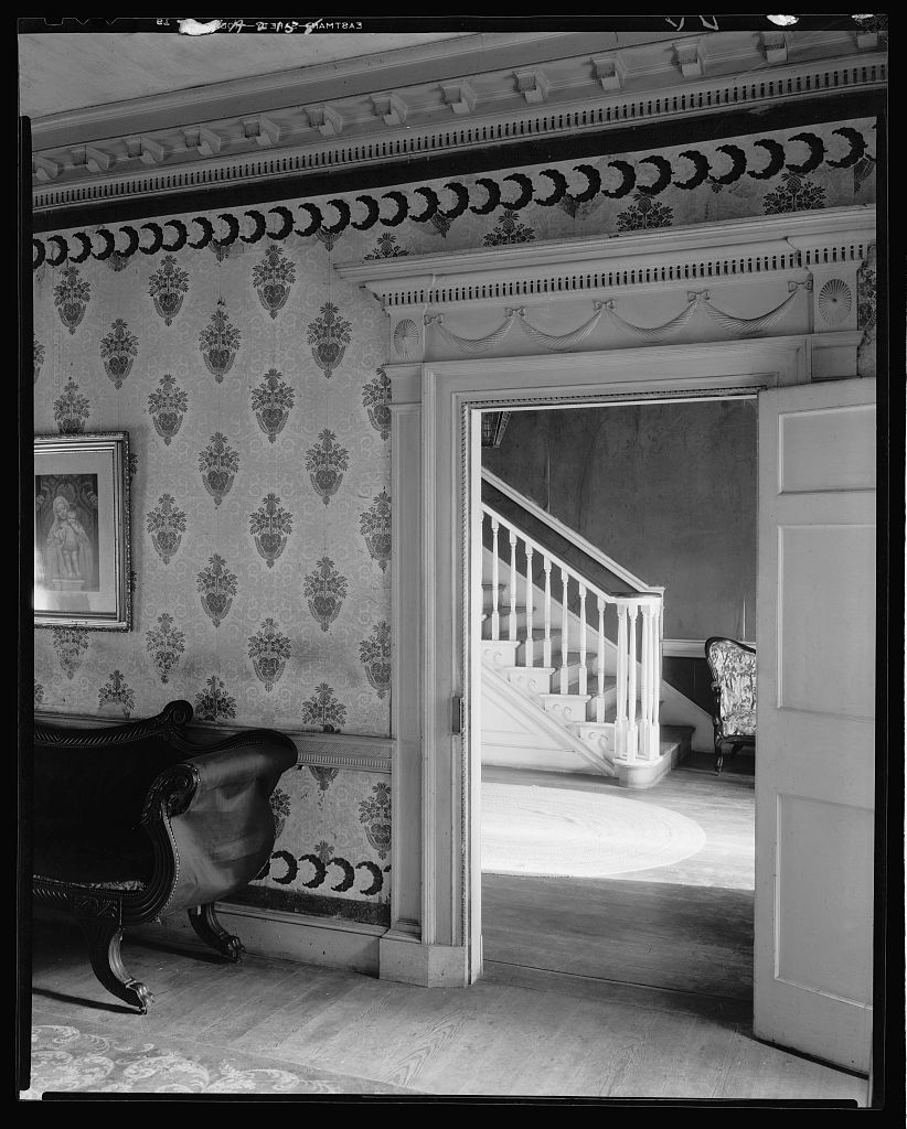 A viewpoint from the inside of a home, depicting a staircase through a doorway, in the foreground a room containing a daybed. 