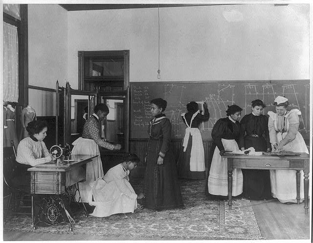 A group of young women Hampton students in a classroom dressmaking. 