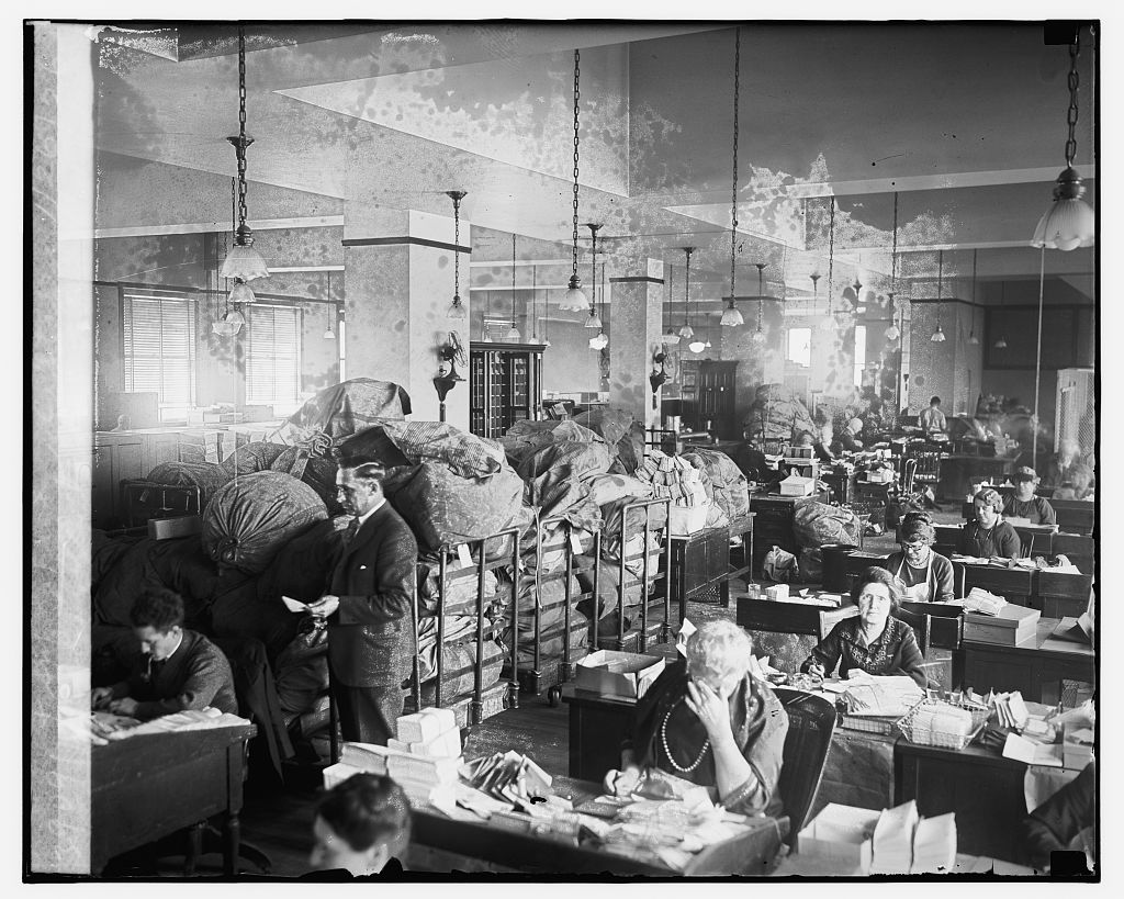 Employees sitting in Dead Letter office surrounded by piles of mail and mail bags. (Source: Library of Congress)