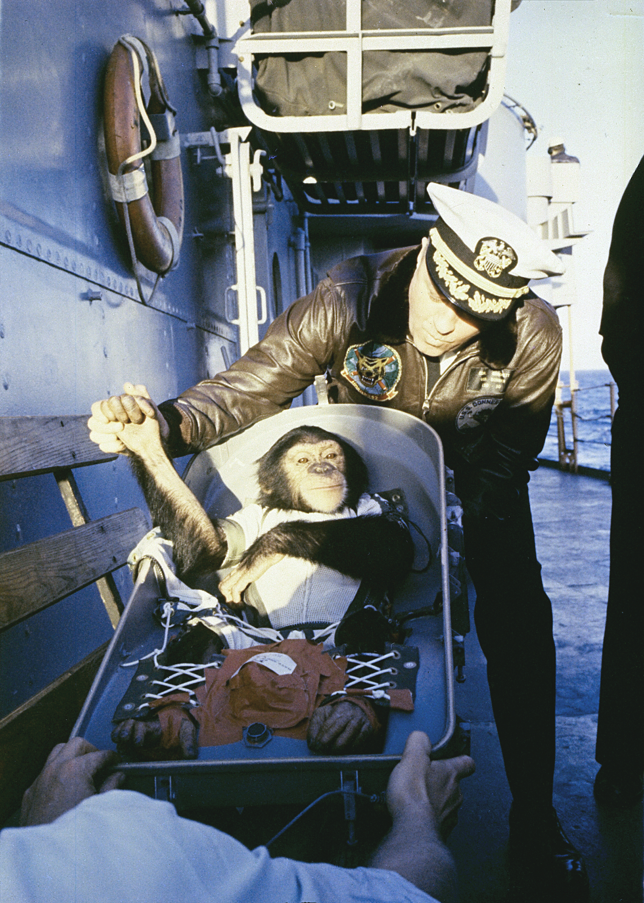 Color photo of a Naval commander standing on the exterior of his ship, warmly grabbing the hand of a young chimpanzee strapped into a capsule.