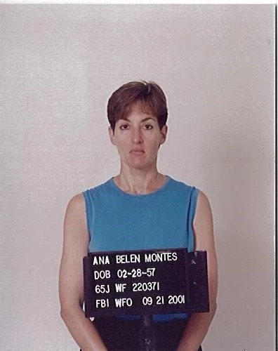 Mugshot of frowning woman with short hair and blue shirt holding a placard with her name, date of birth and FBI ID numbers.