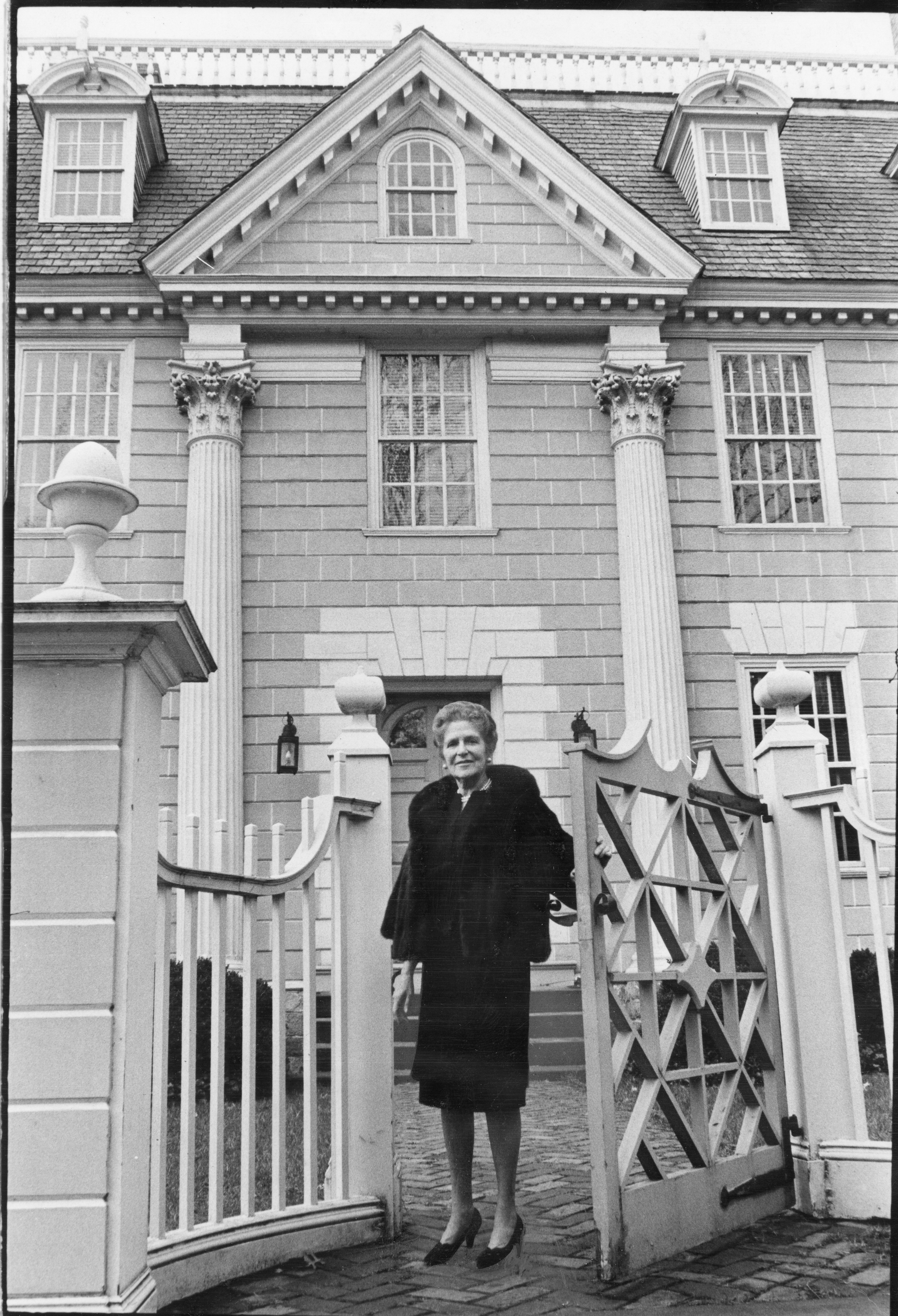A woman in a black dress is standing in front of a house. She is framed by a gate on either side of her.