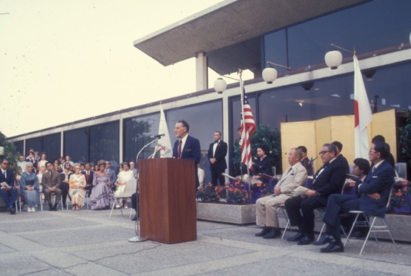 Photo of American man speaking at podium in front of building with American and Japanese flags, and American and Japanese politicians seated behind him, including Secretary of State Henry Kissinger.