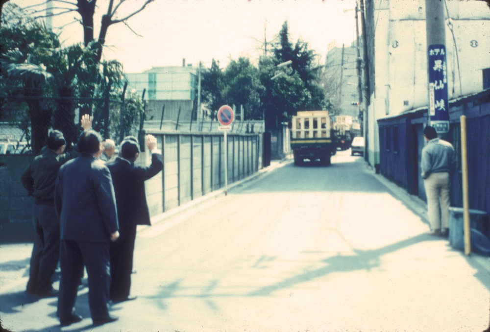 Photo of group of Japanese men in business attire waving goodbye to a truck carrying a crate down a road.