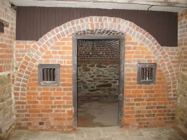 A Photograph of a brick arched entry way with a wooden frame in the middle. Behind the open door is a empty room with a small chandelier hanging from the ceiling