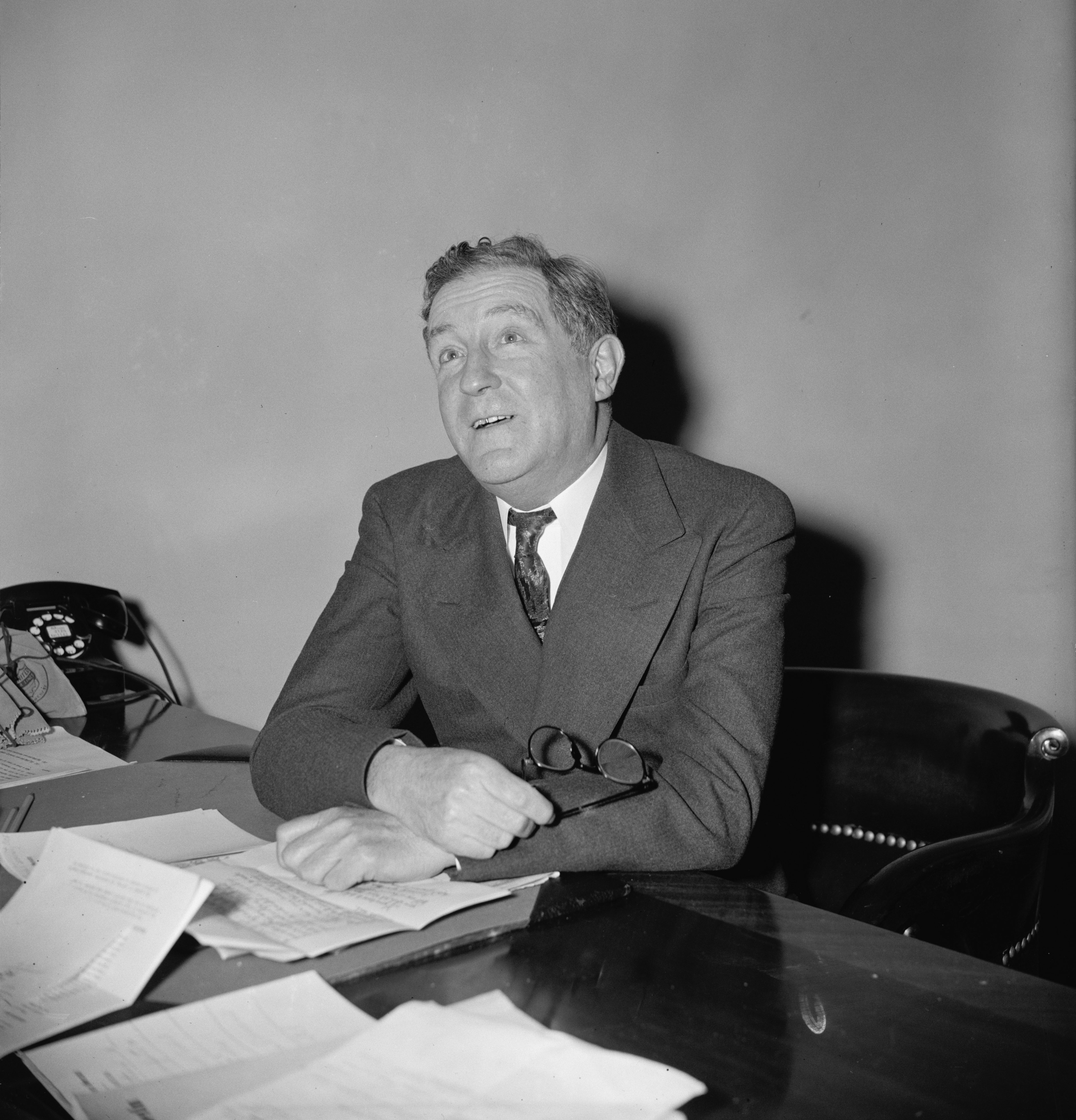 Black and white photo of man sitting at desk covered with papers and looking up toward the ceiling.