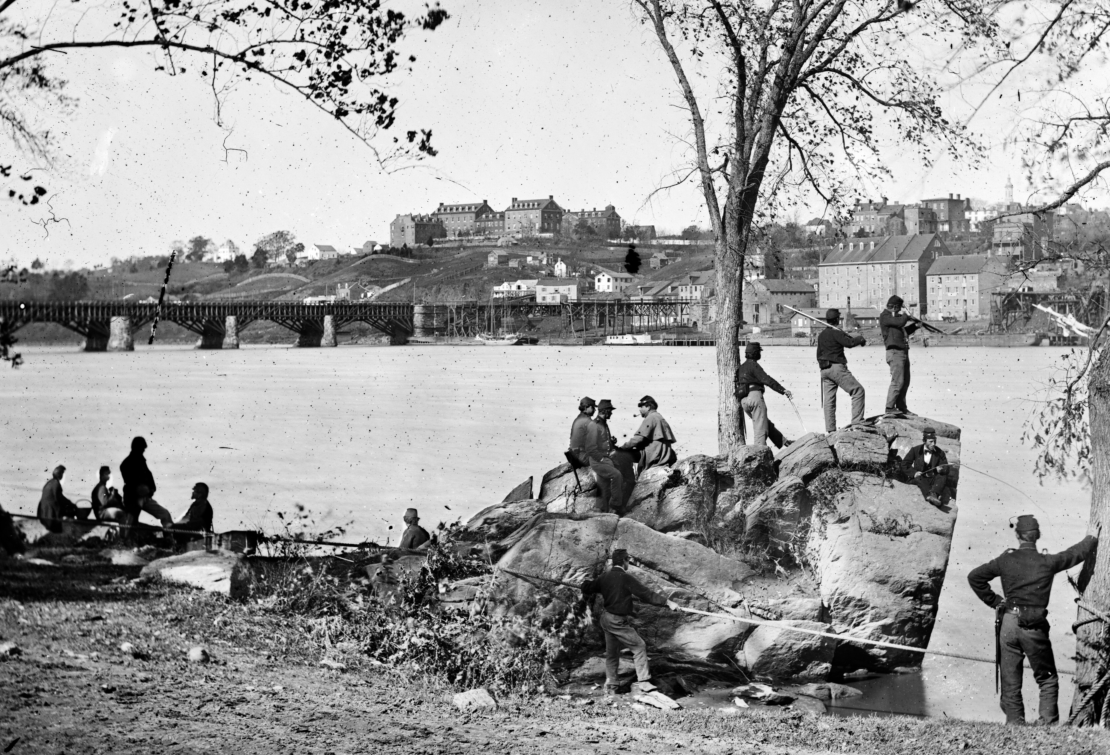 Small group of Civil War soldiers stand near large rocks and look out over Potomac River with Georgetown in the background.