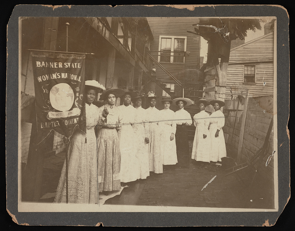 Nine African-American women posed, standing, full length, with Nannie Burroughs holding banner reading, "Banner State Woman's National Baptist Convention"