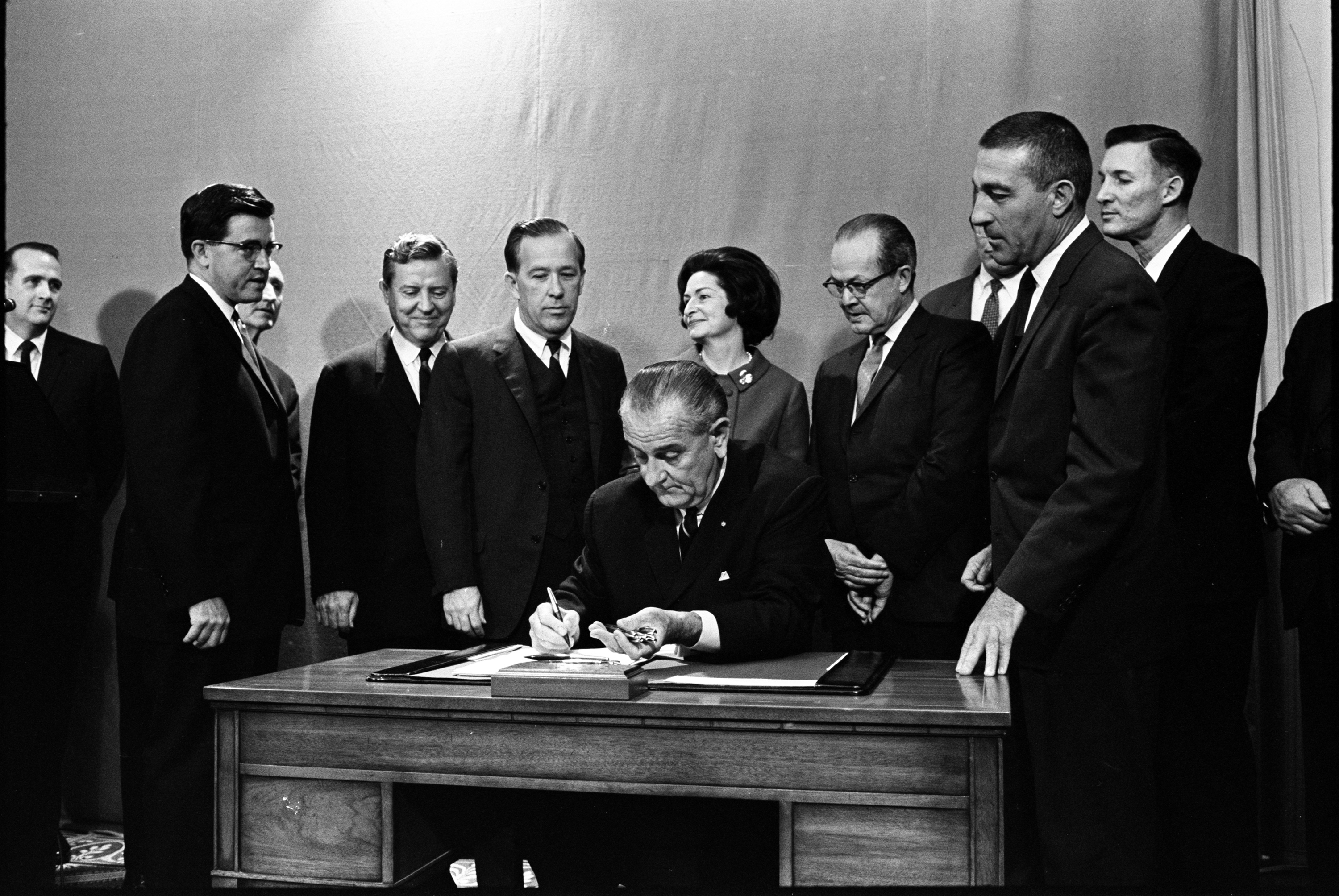 President Lyndon Johnson signs bill as a group of onlookers encircle his desk.