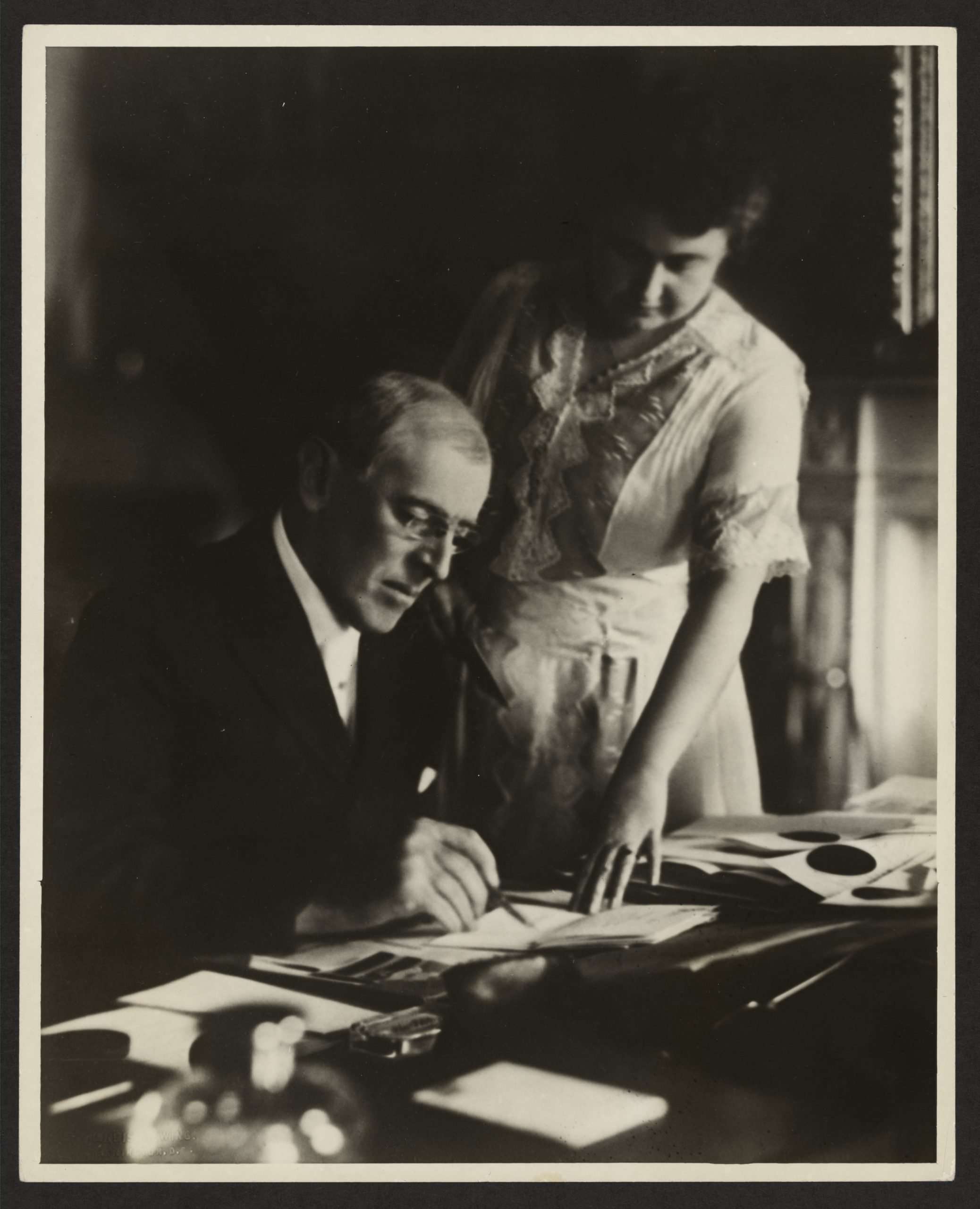 Black and white photo of Edith standing over Woodrow Wilson and working on documents. Source: Encyclopedia Virginia.