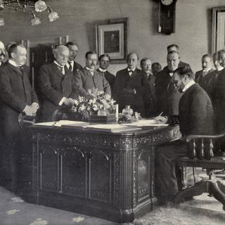 Black-and-white image of John Hay signing the Treaty of Paris, surrounded by other officials.