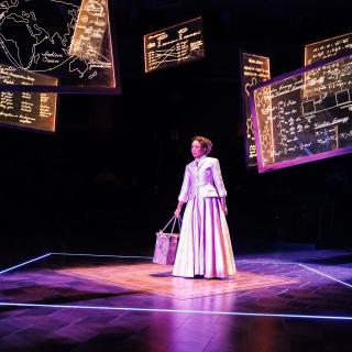 Gina Daniels as Anna Julia Cooper. She stands in a spotlight in the middle of a dark stage wearing a purple suit and holding a bag. Above her are transparent panes of glass with equations, vocabulary, and other lessons written on them