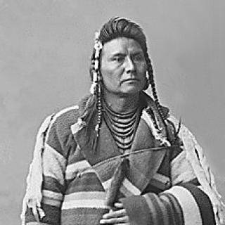 Black and white portrait photo of 39-year-old Chief Joseph standing in front of rock backdrop, wearing striped blanket.