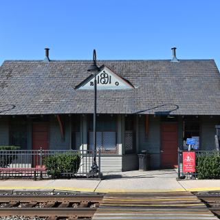 Kensington MARC Station in Maryland, showing a red brick station building with a steeply pitched roof and a prominent tower, alongside train tracks under a partly cloudy sky. A platform with a small shelter is visible, with trees and a few buildings in the background.(Source: G. Edward Johnson on Wikipedia. Cropped and used via Creative Commons Attribution 4.0 International License.)