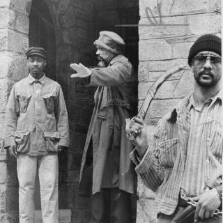 Three men stand in front of stone doorway. One is holding a sword and one is gesturing with his hand.