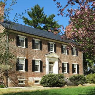 The picture is of a 2 story brick house with a black roof and 4 chimneys, with two on either side. The front of the house is slightly obscured by large bushes that sit along the front and side, and trees which are scattered around the house