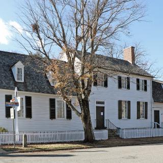 A photo of a large white house with black roof and shutters. The center of the house has a second floor and on either side of it dormer windows are unevenly placed along the roof