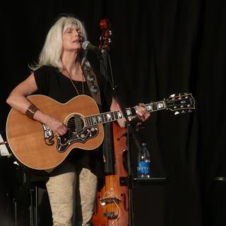 EmmyLou Harris standing, holding a guitar and singing into a microphone.