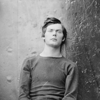 Black and white photo of young man sitting against a wall and wearing handcuffs.