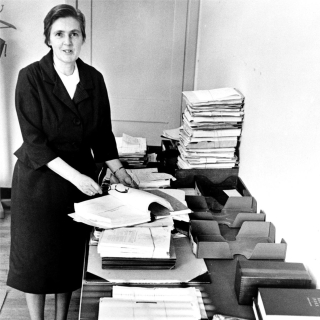 Dr. Frances O. Kelsey, full-length portrait, standing, facing front, in her office at the Food and Drug Administration, next to table with reports on new drugs she has been unable to process since her involvement in refusing to license the drug Thalidomide