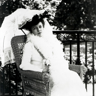 Black and white photo of young woman in white dress and white parasol sitting in chair.