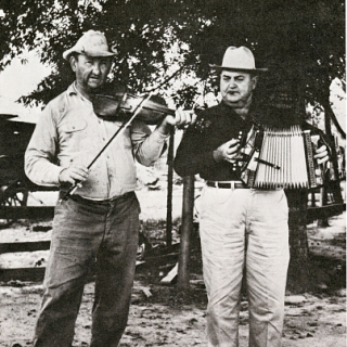Two men standing in the foreground. The left person is playing a violin while the right person is playing an accordion. 