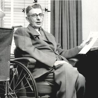 A black-and-white photo. Hugo Deffner, a white man in his 60s, sits in his wheelchair at a desk. He is holding a piece of paper in one hand and wearing a suit. He is looking at the camera.