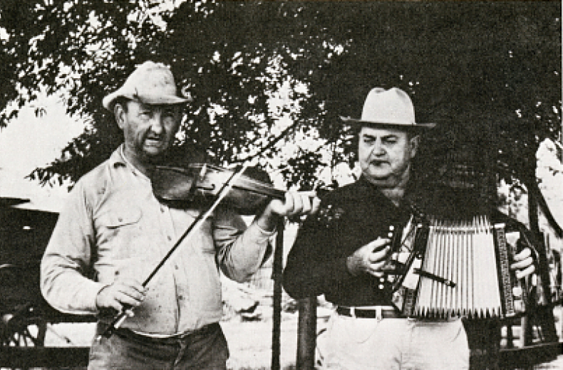 Two men standing in the foreground. The left person is playing a violin while the right person is playing an accordion. 