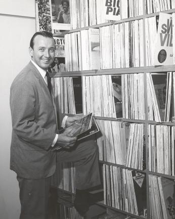 Grant stands beside a row of bookshelves overflowing with records. He holds a few records in his hands. The photo is black and white.