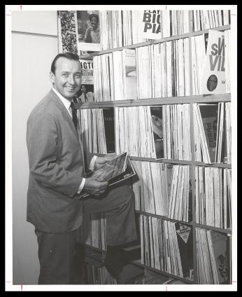 Grant stands beside a row of bookshelves overflowing with records. He holds a few records in his hands. The photo is black and white.