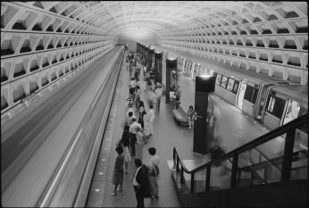 A black-and-white photo of the Foggy Bottom Metro stop. A large group of people wait on one side of the platform while a train waits on the other side with open doors.