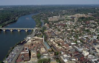 An arial view of Georgetown and the Potomac. On the right side of the photo are crowded buildings, to the left is the river and a bridge, and to the left of the river the land is covered with trees.
