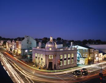 A timelapse photo of the intersection of Wisconsin and M Street in the evening. Car lights create a laser-like effect on the road. The buildings are lit from inside the windows. In the forefront of the photo is the National Bank building