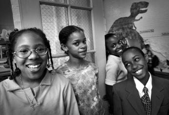 Four Smothers Elementary students smile in a classroom with a t-rex like dinosaur drawn on a wall in the background.