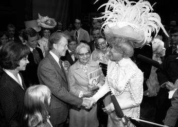 Jimmy Carter shakes actress Carol Channing's hand. They are both grinning widely, Channing is wearing an elaborate white costume, topped with a great hat spewing white feathers in all directions.