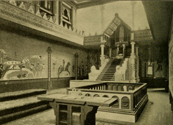 A photo of the Assyrian throne room in the Hall of the Ancients. In view is a large throne and canopy with a model seated in it. Over the walls are paintings