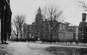 A monochrome photograph featuring the Anne Arundel County courthouse in the background. A path curves in the foreground. The dome is visible behind a screen of tree branches.