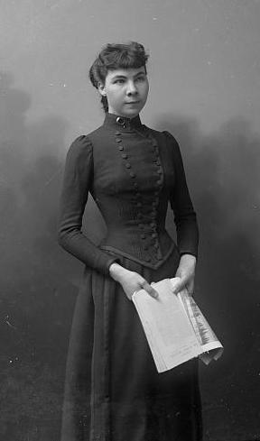 A 3/4 length photo of Madeline Pollard standing in a black dress with a newspaper in her hands