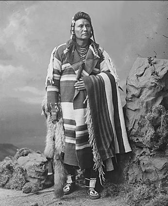 Black and white portrait photo of 39-year-old Chief Joseph standing in front of rock backdrop, wearing striped blanket.