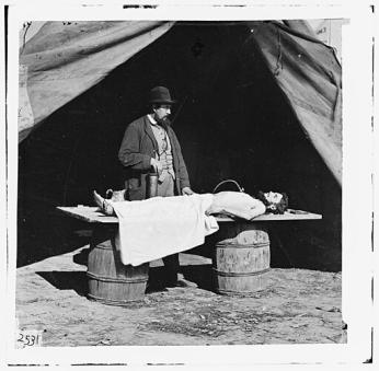 Black-and-white image. In front of a tent, a surgeon in a top hat holds a tube which feeds into the body of a deceased soldier on the table in front of him. (Civil War Glass Negatives, Public domain, via Wikimedia Commons)