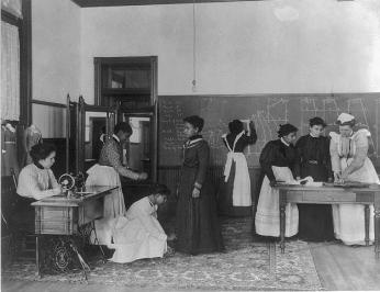 A group of young women Hampton students in a classroom dressmaking. 