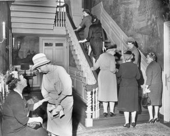 A group of women are talking in a grand hallway lit with candles. Some are going up the stairs.