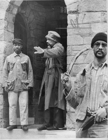 Three men stand in front of stone doorway. One is holding a sword and one is gesturing with his hand.