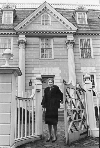 A woman in a black dress is standing in front of a house. She is framed by a gate on either side of her.