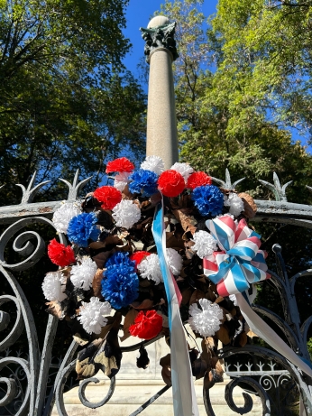 Tall cylindrical monument surrounded by wrought iron fence adorned with red, white and blue flowers.